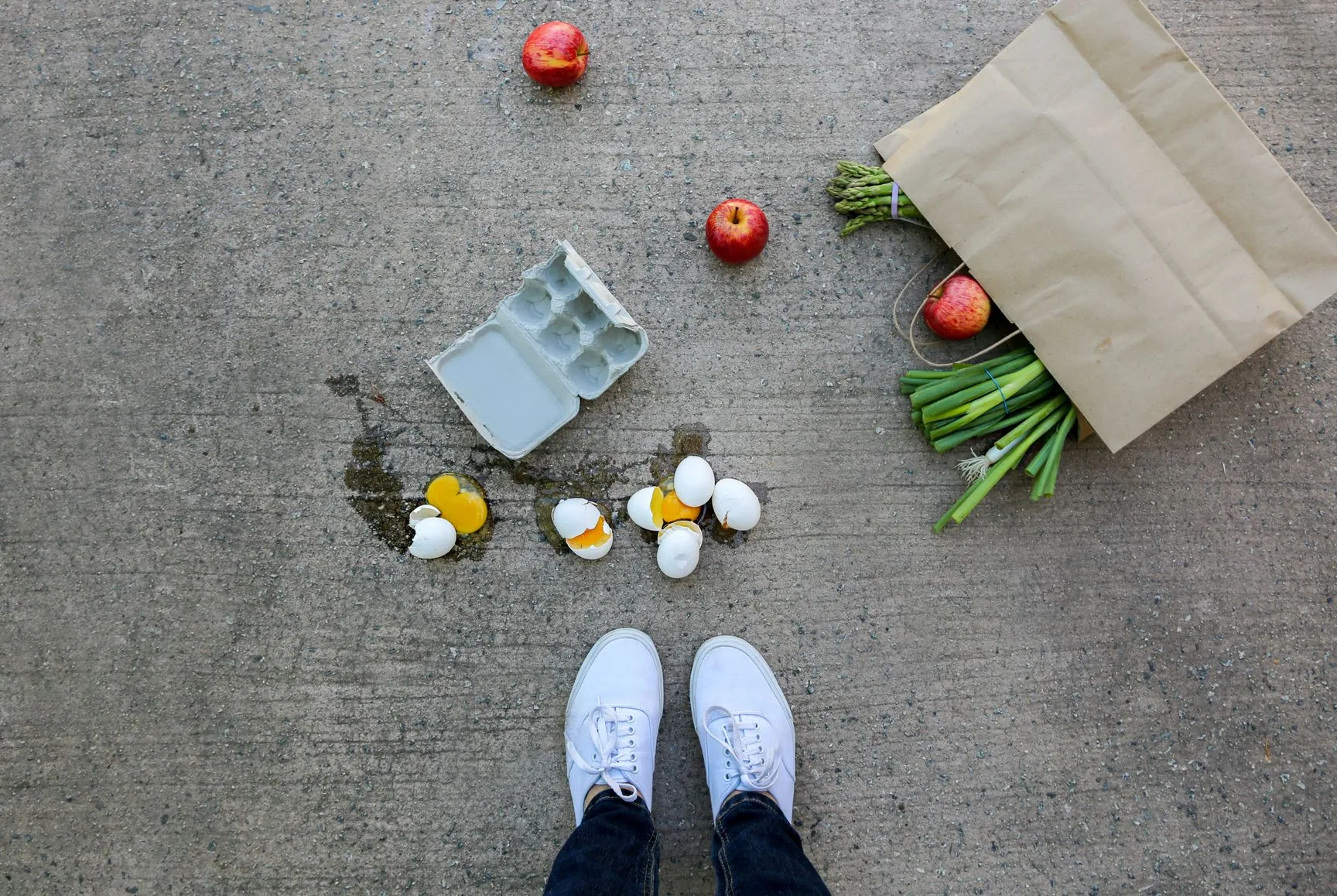 Foto einer heruntergefallenen Papiertüte, aus der Lebensmittel wie Äpfel kullern, daneben eine Packung zerbrochene Hühnereier und die Füße der Person, die das Foto schießt.