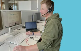 Moritz sitzt an seinem Lieblingsschreibtisch im Office und telefoniert per Headset mit Kollege.