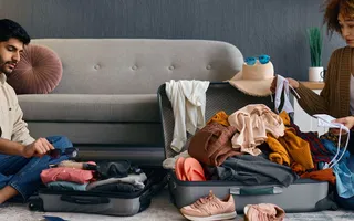 Ein Paar sitzt vor dem Sofa auf dem Boden und packt (recht chaotisch) seine Siebensachen für den Urlaub.