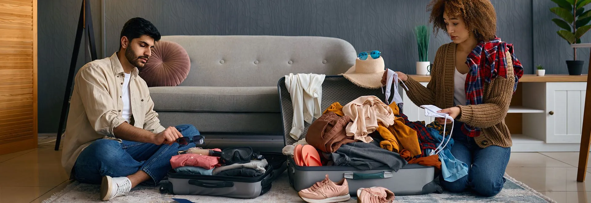 Ein Paar sitzt vor dem Sofa auf dem Boden und packt (recht chaotisch) seine Siebensachen für den Urlaub.