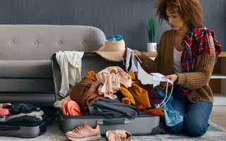 Ein Paar sitzt vor dem Sofa auf dem Boden und packt (recht chaotisch) seine Siebensachen für den Urlaub.