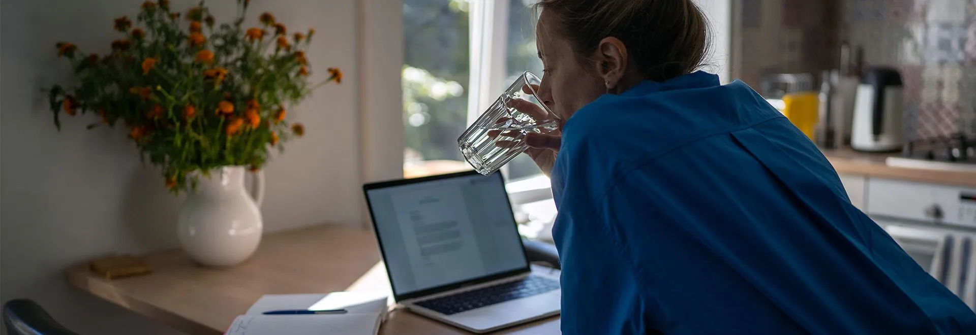 Eine Frau im ultramarinblauen Hemd lehnt am Küchentisch und trinkt ein Glas Wasser, während sie auf den Laptop und Schreibutensilien vor sich blickt. Ob sie gerade über Obliegenheiten in ihrer Versicherungen nachdenkt?