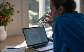 Eine Frau im ultramarinblauen Hemd lehnt am Küchentisch und trinkt ein Glas Wasser, während sie auf den Laptop und Schreibutensilien vor sich blickt. Ob sie gerade über Obliegenheiten in ihrer Versicherungen nachdenkt?