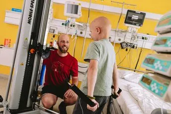 Ein Sporttherapeut behandelt einen kleinen Patienten an einer Sportmaschine in einem gelben Krankenhauszimmer.