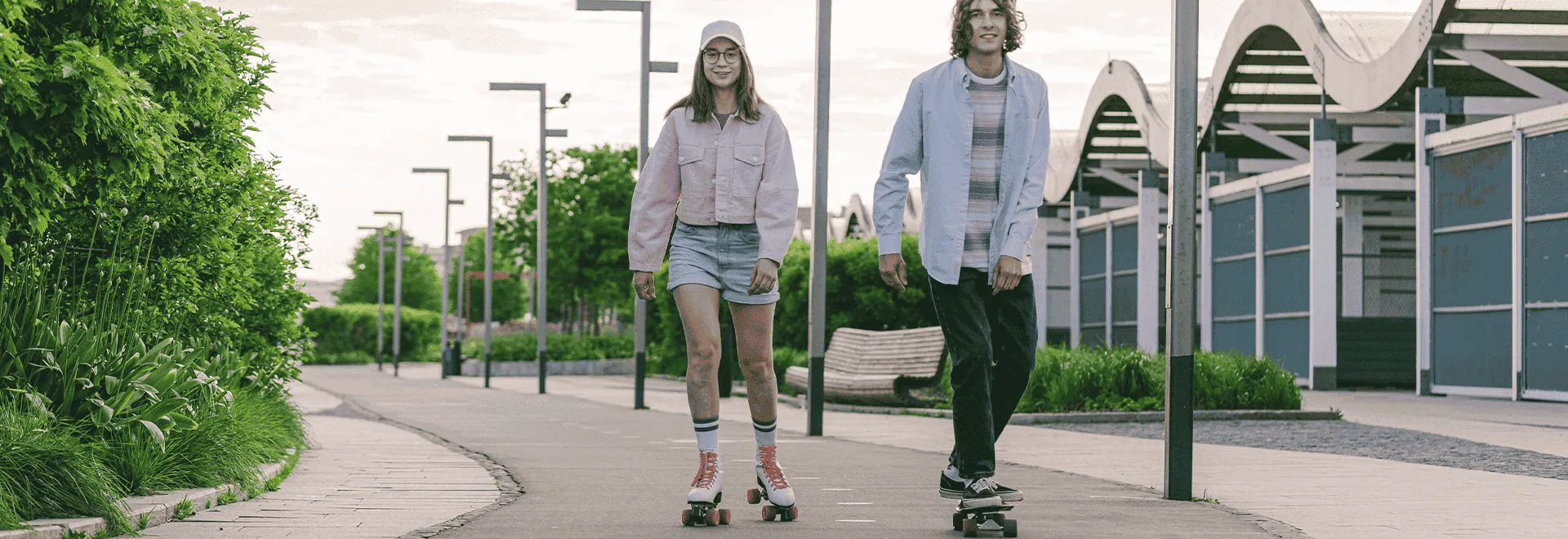 Für Aktive lohnt sich eine Unfallversicherung häufig: eine Rollerskaterin und ein Skateboarder fahren auf einer asphaltierten Promenade.