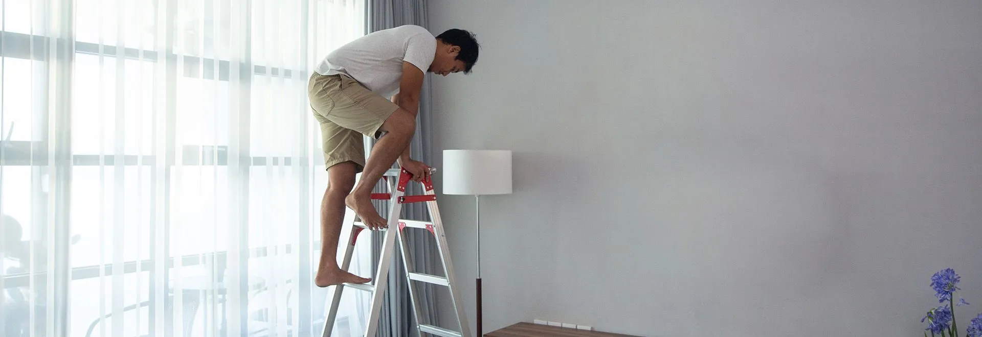 Häuslicher Unfall am entstehen: Ein Mann klettert barfuß auf einer Leiter im Wohnzimmer, um eine Blühbirne zu wechseln.