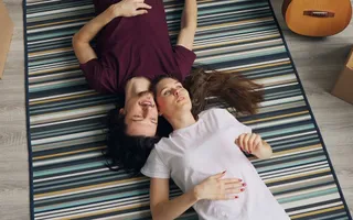 Ein Paar liegt in ihrem neuen Zuhause auf dem frisch ausgerollten Teppich; um sie herum Umzugskisten. Vielleicht fragen sie sich gerade: Was kostet eine Wohngebäudeversicherung?