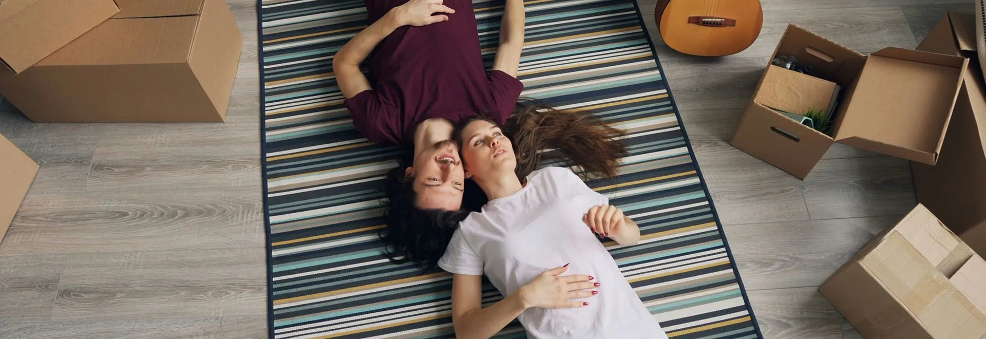 Ein Paar liegt in ihrem neuen Zuhause auf dem frisch ausgerollten Teppich; um sie herum Umzugskisten. Vielleicht fragen sie sich gerade: Was kostet eine Wohngebäudeversicherung?