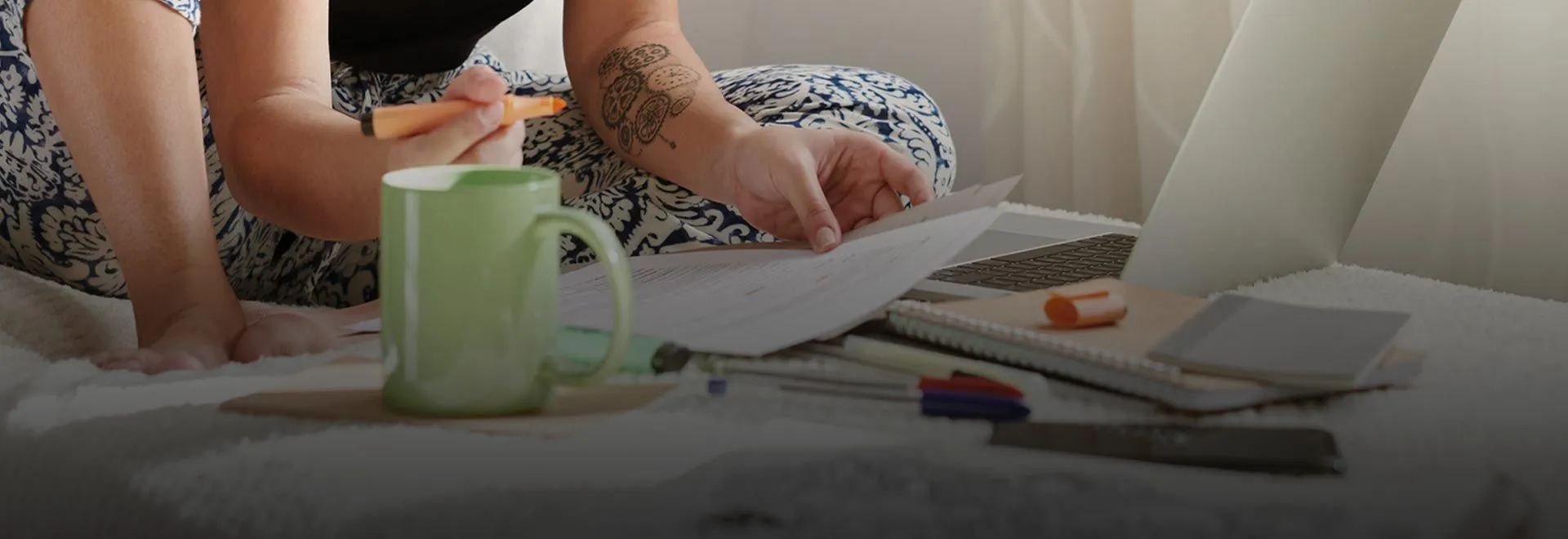 Eine Frau sitzt auf einem Bett mit einem Laptop und anderen Büroartikeln und hält Dokumente in der Hand.