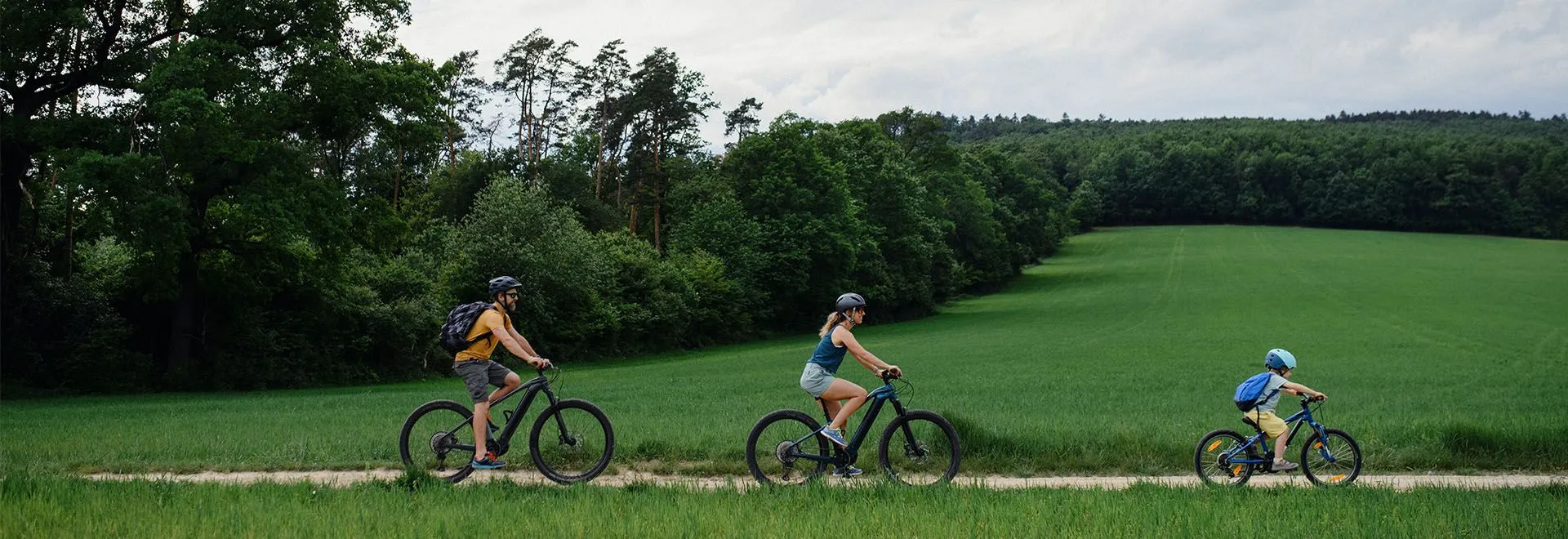 Eine Familie mit Mutter, Vater und Kind fährt in kurzen Outfits E-Bike, im Hintergrund sieht man ein sommerlich grünes Feld und Waldrand.