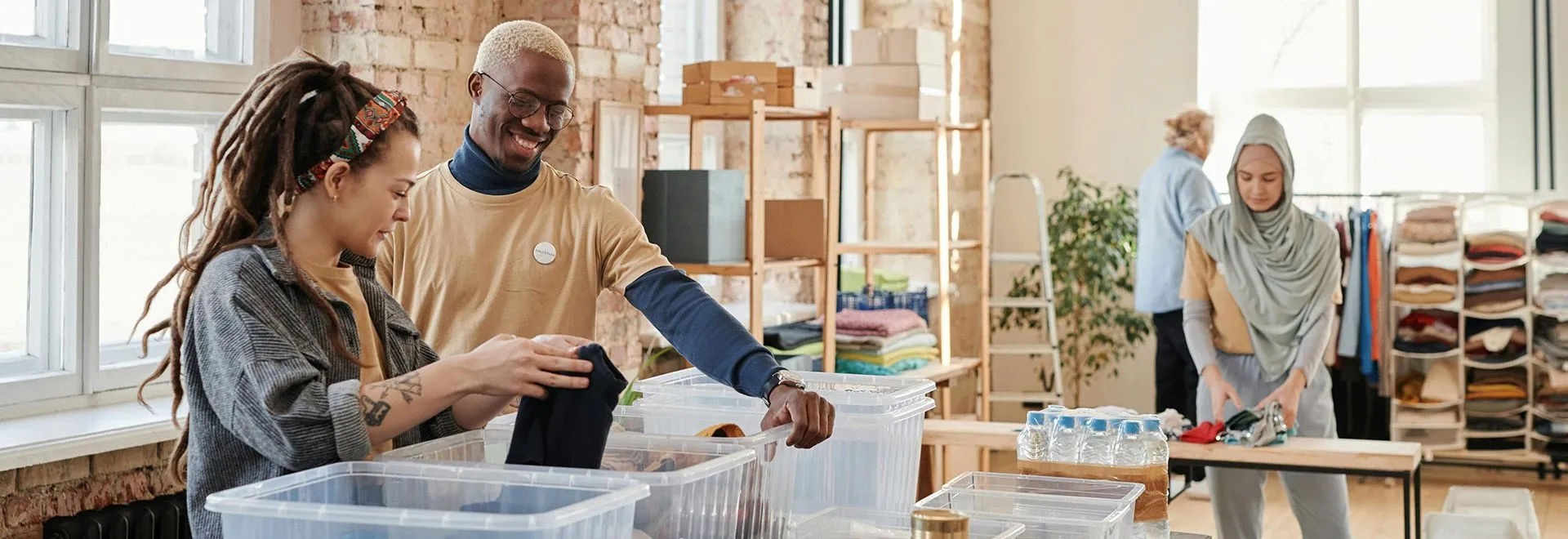 Drei Ehrenamtliche sortieren Spenden wie Kleidung und Getränke in transparente Boxen.