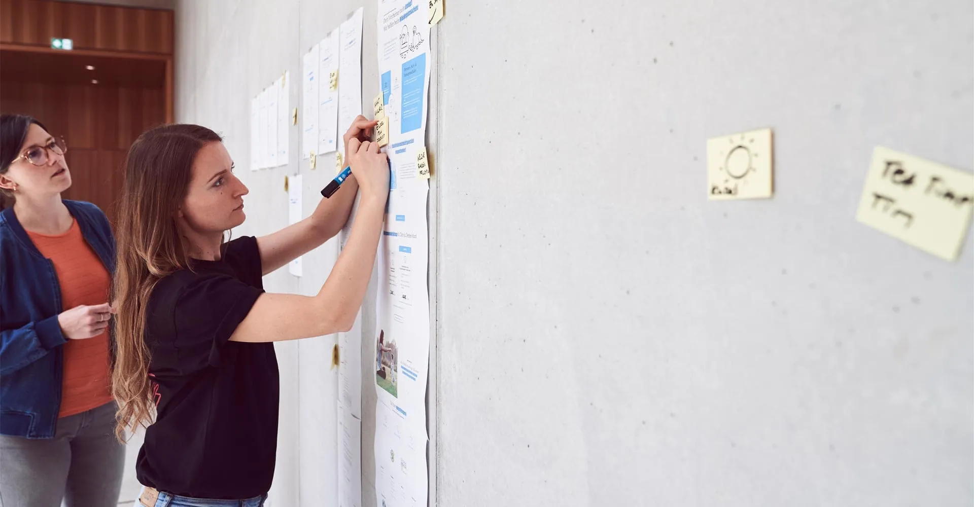 Janette mit einer Kollegin, wie sie auf Post-its an einer Pinnwand schreiben