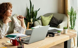 Frau im Homeoffice, die am Schreibtisch vor ihrem Laptop sitzt und Richtung Fenster schaut. 