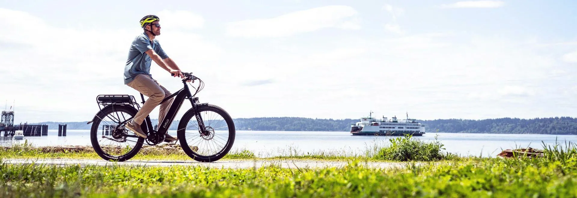 Ein Mann fährt E-Bike auf einer frühlingshaften Wiese entlang eines Flusses. Die Sonne scheint und im Himmel hängen ein paar Schönwetterwolken. Im Hintergrund sieht man ein Schiff den Fluss hinabfahren.
