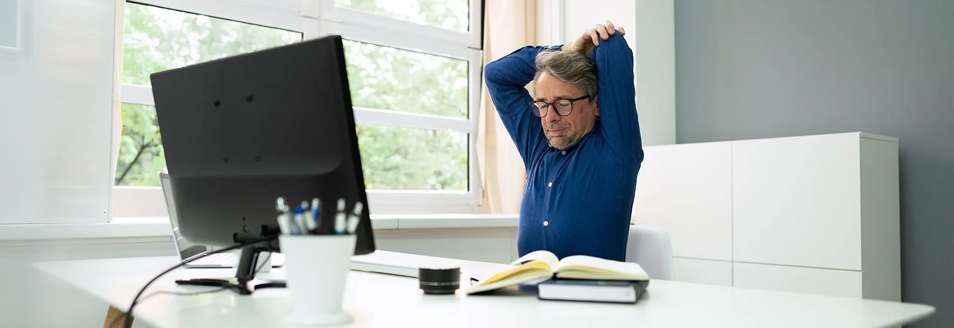 Ein Mann im blauen Hemd sitzt an seinem Schreibtisch und macht Dehungsübungen.