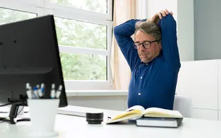 Ein Mann im blauen Hemd sitzt an seinem Schreibtisch und macht Dehungsübungen.