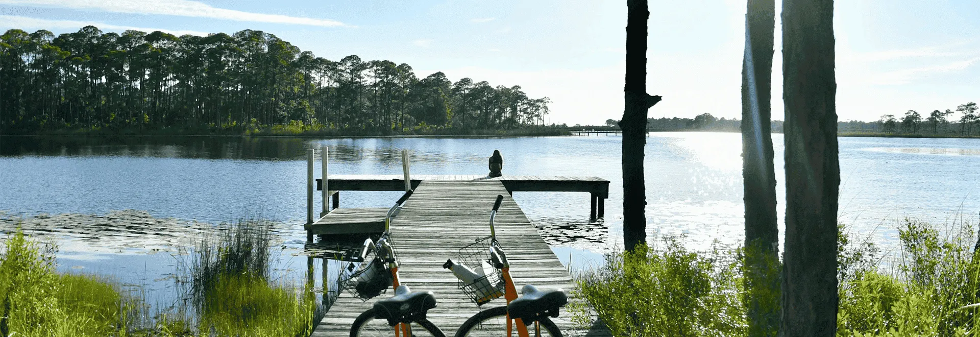 Lohnt sich eine E-Bike-Versicherung auf einem Ausflug? Zwei E-Bikes stehen vor einem Steg, auf dem eine Frau sitzt und auf den See schaut. Im Hintergrund sieht man einen Kiefernwald.