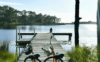 Lohnt sich eine E-Bike-Versicherung auf einem Ausflug? Zwei E-Bikes stehen vor einem Steg, auf dem eine Frau sitzt und auf den See schaut. Im Hintergrund sieht man einen Kiefernwald.