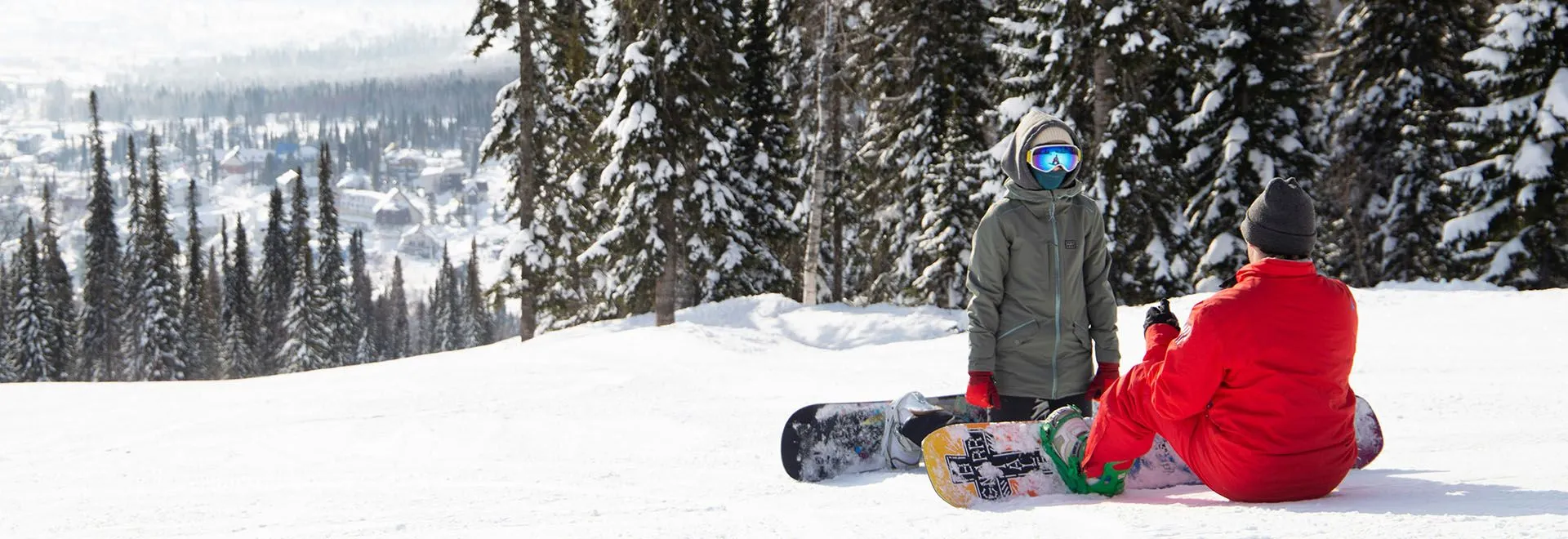 Zwei Snowboarder knien im Schnee am Rande einer Piste, im Hintergrund Wald.