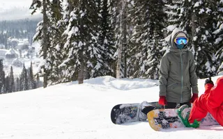 Zwei Snowboarder knien im Schnee am Rande einer Piste, im Hintergrund Wald.