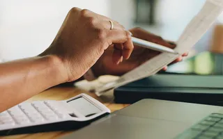 Nahaufnahme einer Person am Schreibtisch mit Papieren, Laptop und Taschenrechner. 