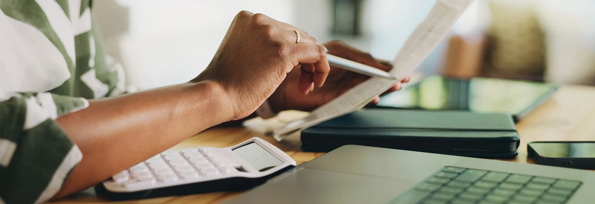 Nahaufnahme einer Person am Schreibtisch mit Papieren, Laptop und Taschenrechner. 
