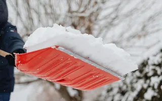 Ein Mann in dicker Winterjacke schippt Schnee, im Vordergrund sieht man die erhobene rote Schneeschaufel, im Hintergrund Hecke und ein Baum.