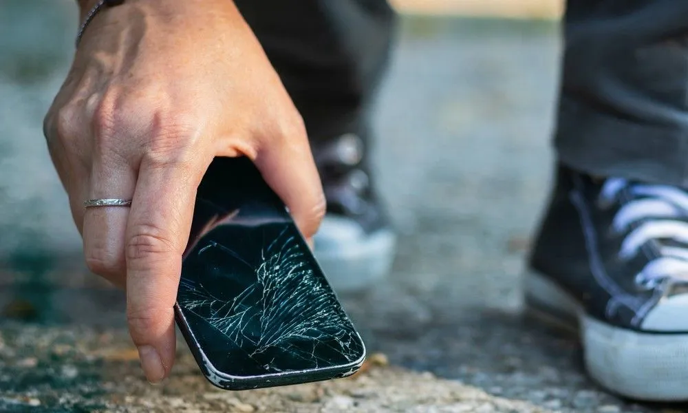 Eine Person hebt ein Handy mit gesprungenem Display vom Boden auf.