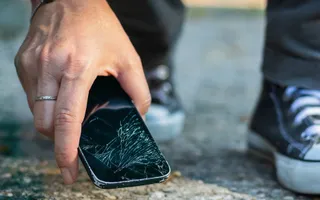 Eine Person hebt ein Handy mit gesprungenem Display vom Boden auf.