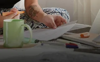 Eine Frau sitzt auf einem Bett mit einem Laptop und anderen Büroartikeln und hält Dokumente in der Hand.