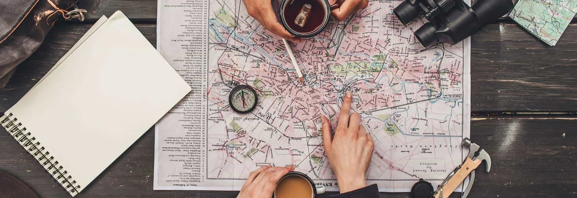 Eine Landkarte und weitere Reiseutensilien liegen auf einem Tisch, eine Hand zeigt auf einen Punkt auf der Karte.