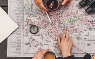 Eine Landkarte und weitere Reiseutensilien liegen auf einem Tisch, eine Hand zeigt auf einen Punkt auf der Karte.