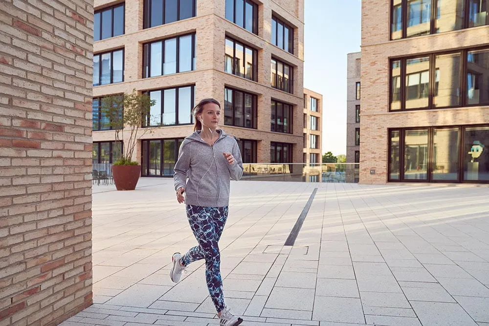 Janette joggt zwischen Bürogebäuden in blau gemusterten Leggings und einer grauen Sportjacke.