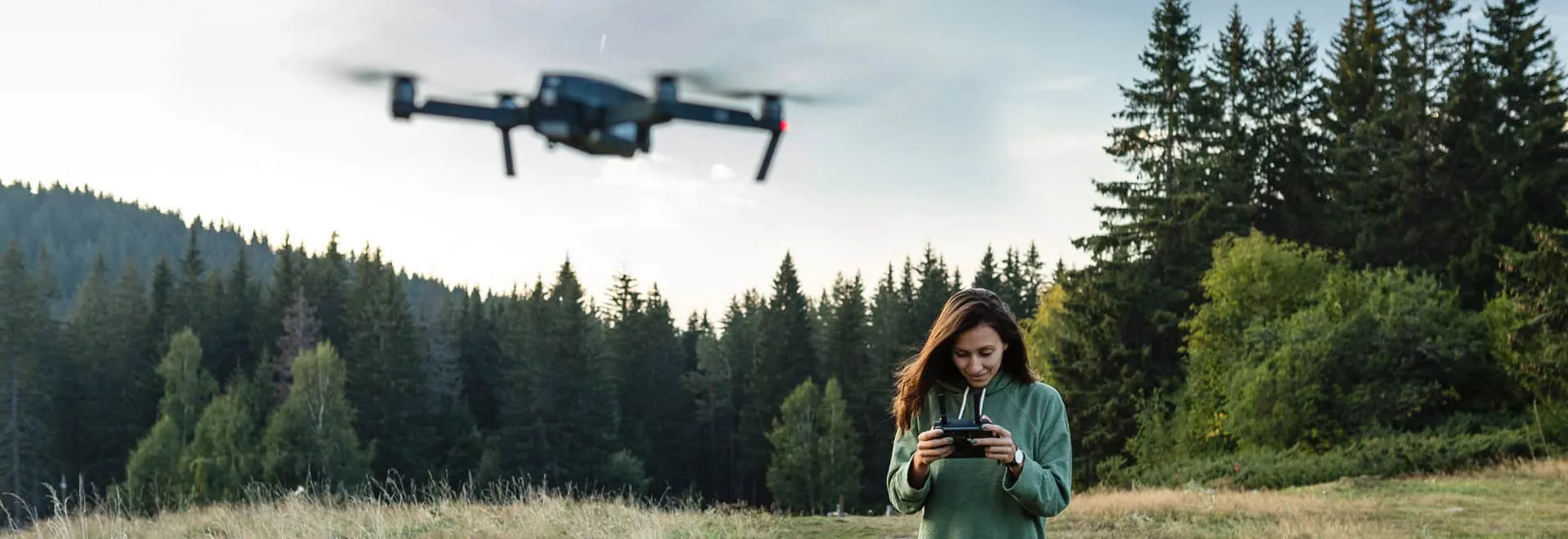 Frau steuert Drohne auf einer großen Wiese vor Wald